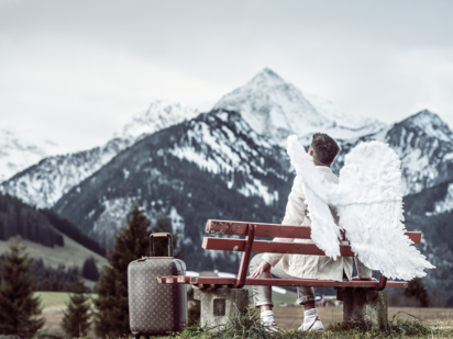 Ein Engel auf einer roten Bank im Tannheimer Tal mit Koffer bei der Anreise | Der Engel in Tirol