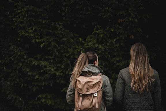 Zwei Personen in grünen Jacken stehen vor grünen Bäumen mit traditionellem Holzzaun im Hintergrund