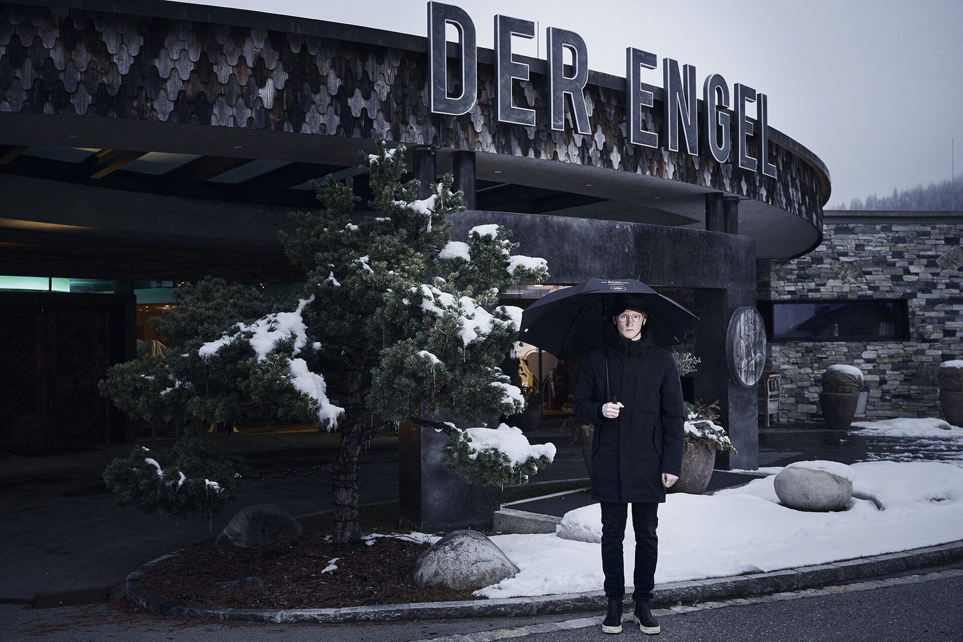 Mann steht im Winter vor dem Eingang zum Hotel der Engel mit Regenschirm in schwarzem Outfit | Der Engel in Tirol