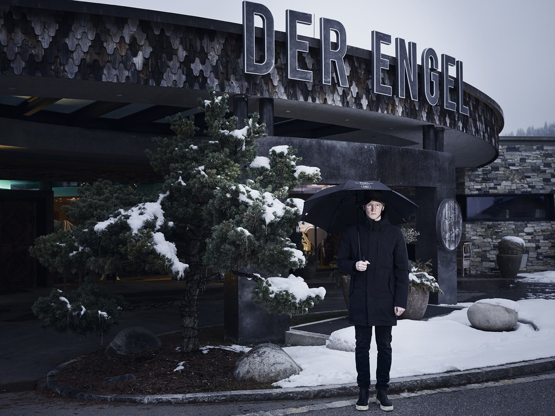 Mann steht im Winter vor dem Eingang zum Hotel der Engel mit Regenschirm in schwarzem Outfit | Der Engel in Tirol
