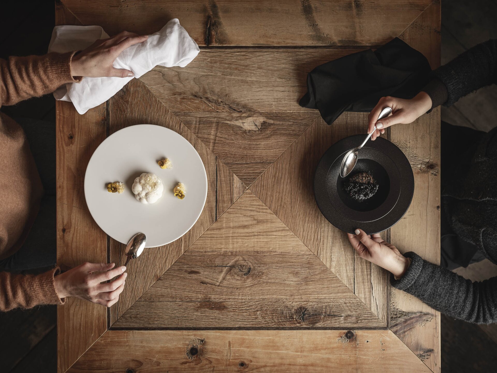 Zwei Personen sitzen an einem alten Holztisch und essen monochrome Gerichte - weißes Gericht auf weißem Teller und schwarzes Gericht auf schwarzem Teller