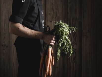 Koch vom Wellnesshotel Der Engel, gekleidet im schwarzen Kochoutfit, hält ein Bündel Karotten am Karottengrün in der Hand
