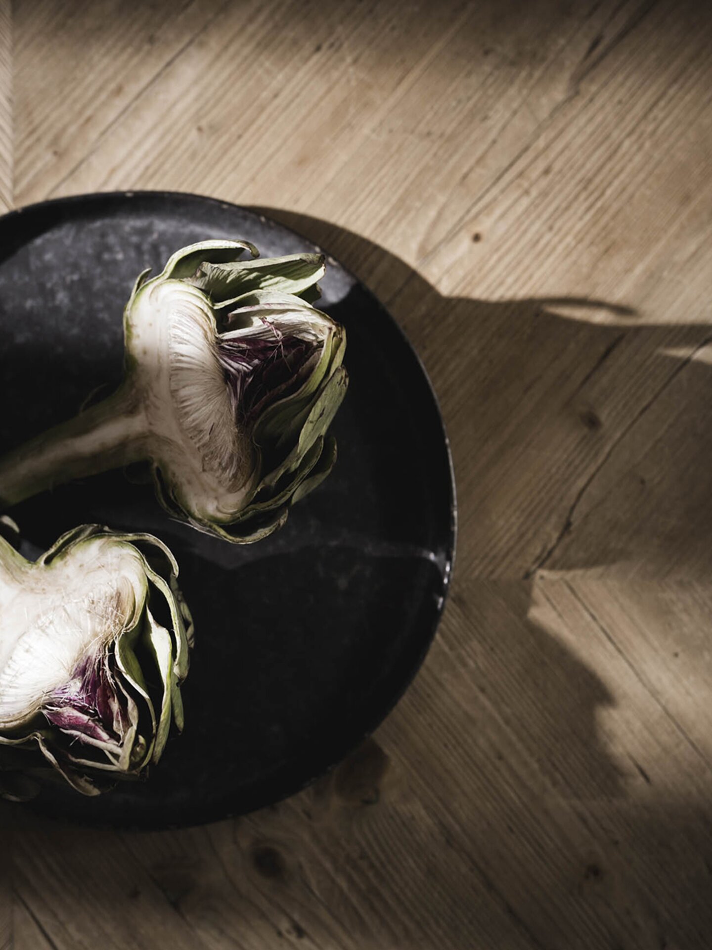 Geteilte Artischocke auf einem schwarzen Teller auf einem Holzuntergrund im Licht und Schatten-Spiel