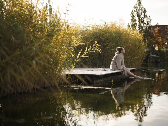 Person sitzt im Bademantel am Holzsteg am Rande des Engel Bergsees zwischen Schilf