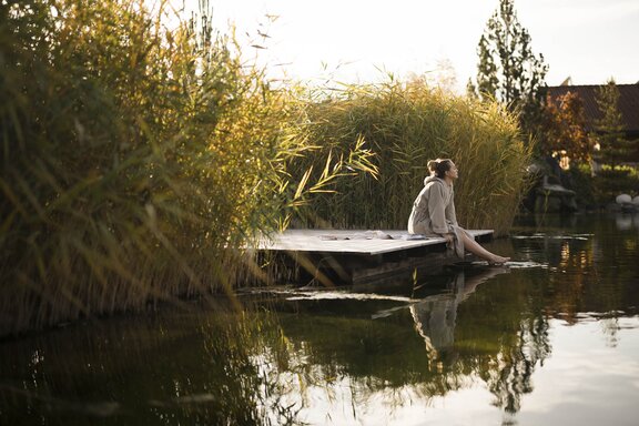 Person sitzt im Bademantel am Holzsteg am Rande des Engel Bergsees zwischen Schilf