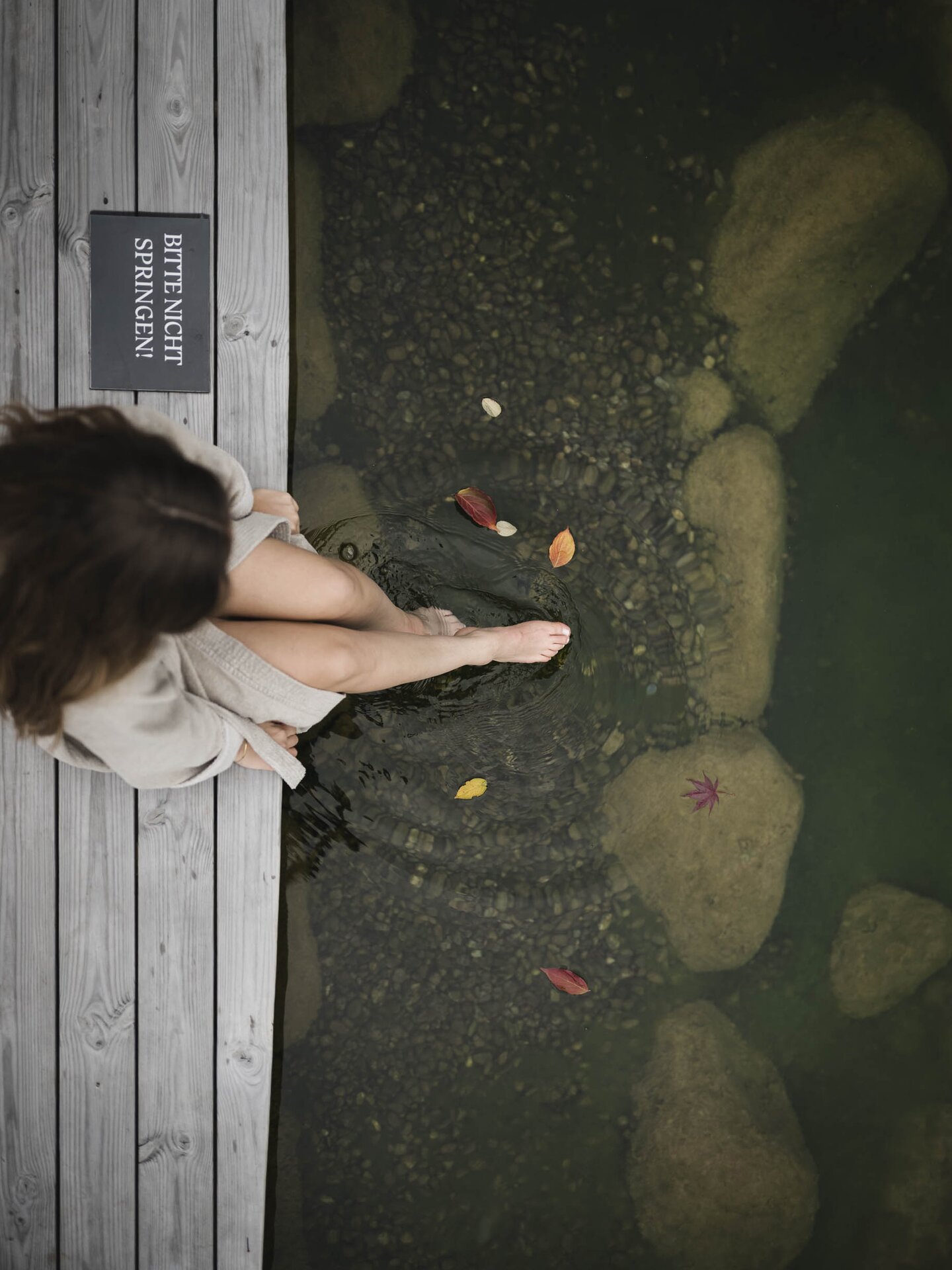 Person sitzt im Bademantel am Holzsteg und lässt die Füße im Wasser baumeln