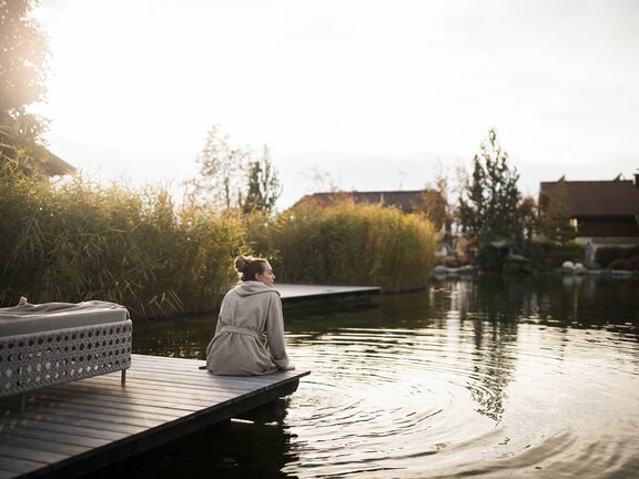 Person sitzt mit Rücken zur Kamera gekehrt im Bademantel auf dem Holzsteg und lässt die Füße im kalten Engel Bergsee baumeln