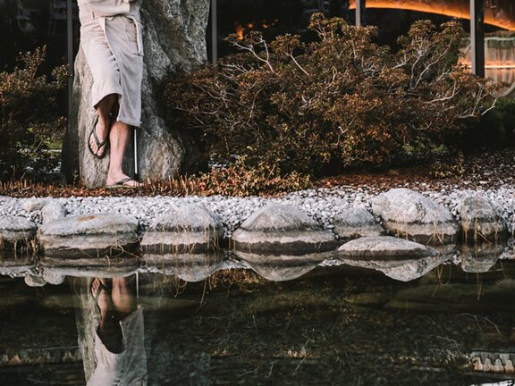 Wellnessgast im Bademantel lehnt an Stein-Dusche am Rande des Bergsees im Herbst im Wellnesshotel Der Engel