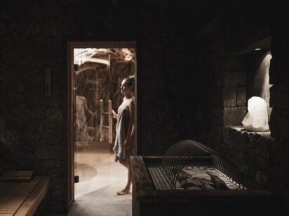 Person, mit Handtuch umwickelt betritt die Steinhaus-Sauna, welche mit dunkelgrauen Natursteinen verkleidet ist