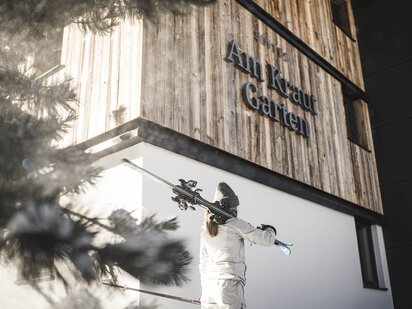 Person im beigen Skioutfit und Skiern auf den Schultern vor verschneiten Suites am Krautgarten