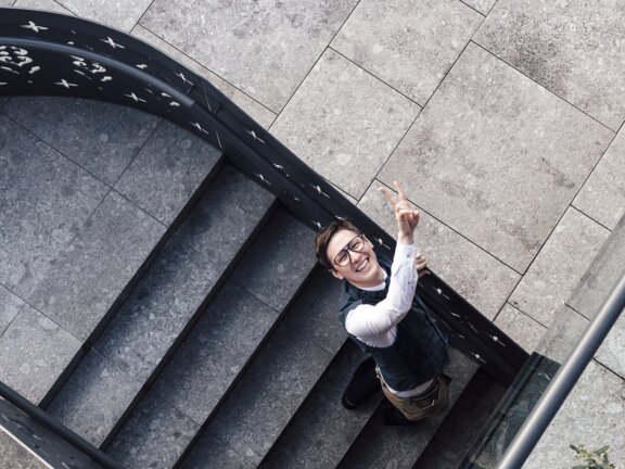 Toni Schlachter - Hoteldirektor - steht auf der Treppe im Außenbereich des Wellnesshotels Der Engel und lächelt in die Kamera und zeigt Peace