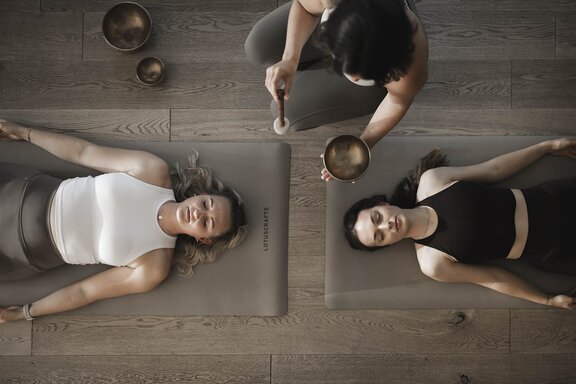 Zwei Frauen liegen auf einer beigen Yogamatte bei einer Klangschalenmeditation im Wellnesshotel Der Engel