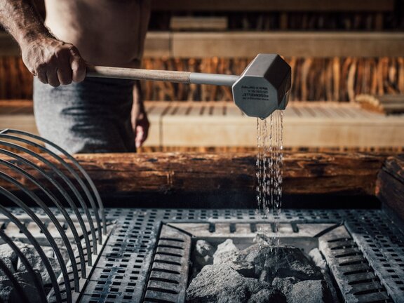 Saunameister beim Saunaufguss, Wasser wird über Saunaofen gegossen | Wellnesshotel Der Engel